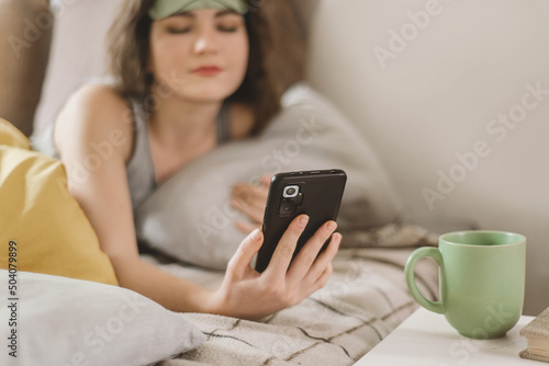 Sleepy young woman in a sleep mask reaches for her smartphone while lying in bed in a sunny bedroom. Turn off the alarm on your smartphone