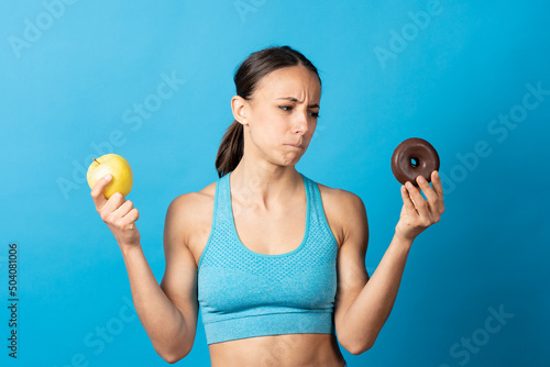 Fotobehang Hispanic fit woman looking at donut with envy and holding apple isolated on blue background