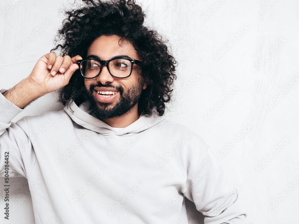 Handsome smiling hipster  model.Sexy unshaven Arabian man dressed in summer stylish hoodie clothes. Fashion male with long curly hairstyle posing near grey wall in studio. Isolated