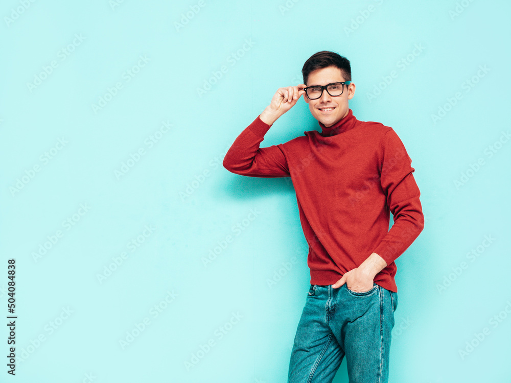 Fototapeta premium Portrait of handsome smiling model. Sexy stylish man dressed in red turtleneck sweater and jeans. Fashion hipster male posing near blue wall in studio. Isolated