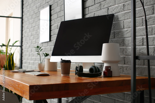 Modern computer, paper cup of drink and headphones on wooden standing desk near grey brick wall