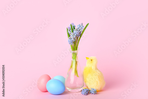 Cute yellow chicken, vase with flowers and Easter eggs on pink background