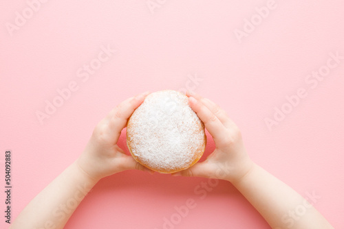 Baby girl hands holding fresh donut with white sprinkled sugar on light pink table background. Pastel color. Closeup. Point of view shot. Sweet snack. Top down view.