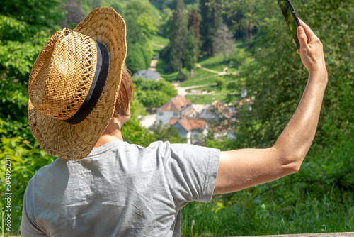 Woman with brown hair, gray t-shirt and straw hat sitting on a park bench in front of Staatspark Fürstenlager, Auerbach, Taking a Selfie with her smartphone, rear view, Bensheim, Germany photo