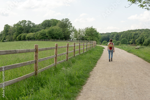 Woman with brown hair, gray t-shirt, jeans and straw hat attached to a red backpack hiking next to a paddock in the nature, rear view © Bildgigant