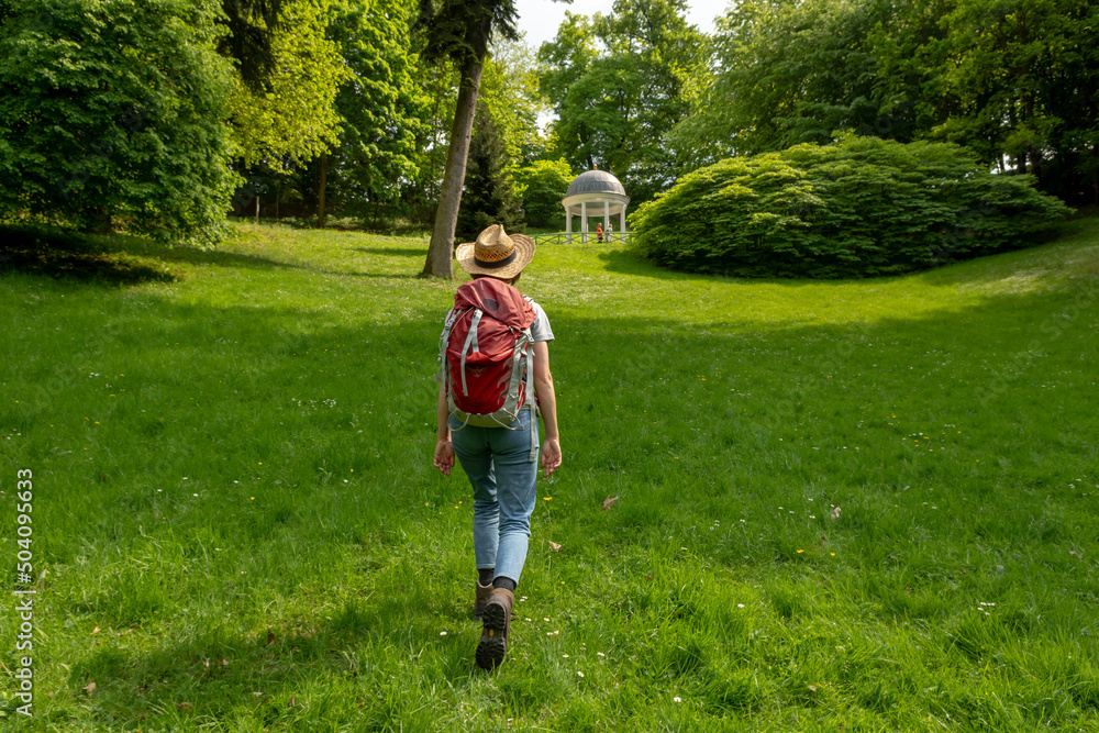 Woman with brown hair, gray t-shirt, jeans, straw hat and red backpack hiking on a meadow at Freundschaftstempel temple, Staatspark Fürstenlager, Auerbach, Bensheim, Germany Woman with brown hair, gray t-shirt, jeans, straw hat and red backpack hiking on a meadow at Freundschaftstempel temple, Staatspark Fürstenlager, Auerbach, Bensheim, Germany