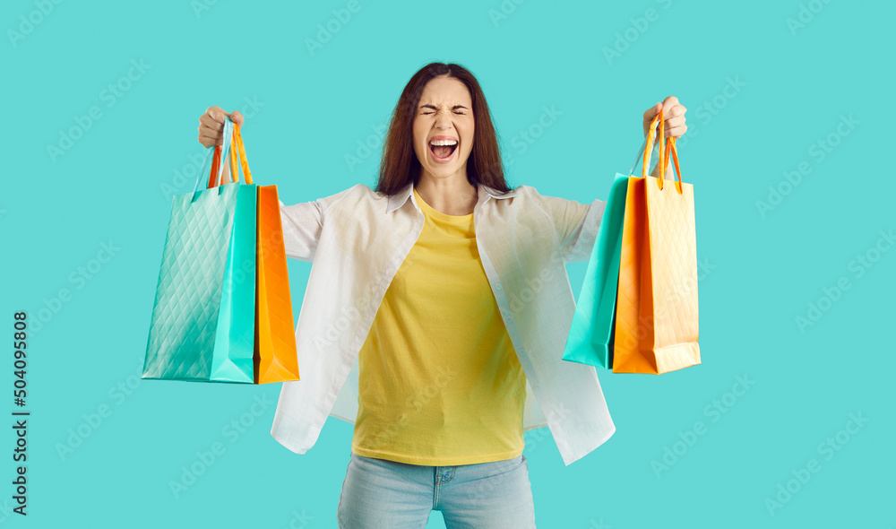Crazy joyful woman with colorful packets shouts excited from her shopping at seasonal sales. Casual woman with closed eyes squeaking loudly while holding shopping bags on light blue background. Banner