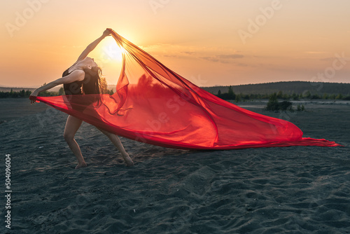 Elegant girl performs dance moves on sand holding up long red fabric against sunset.