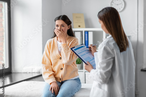 medicine, healthcare and people concept - female doctor with clipboard talking to scared or coughing woman patient at hospital