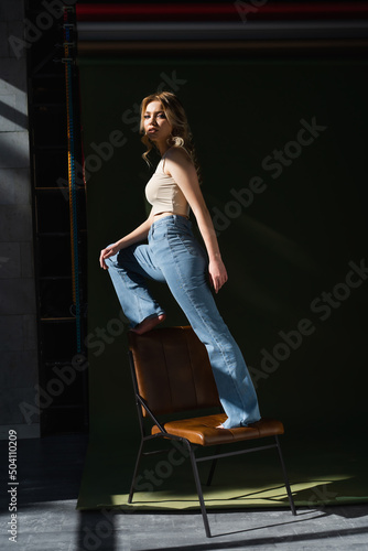 full length of barefoot woman in jeans looking at camera while standing on chair on dark background.