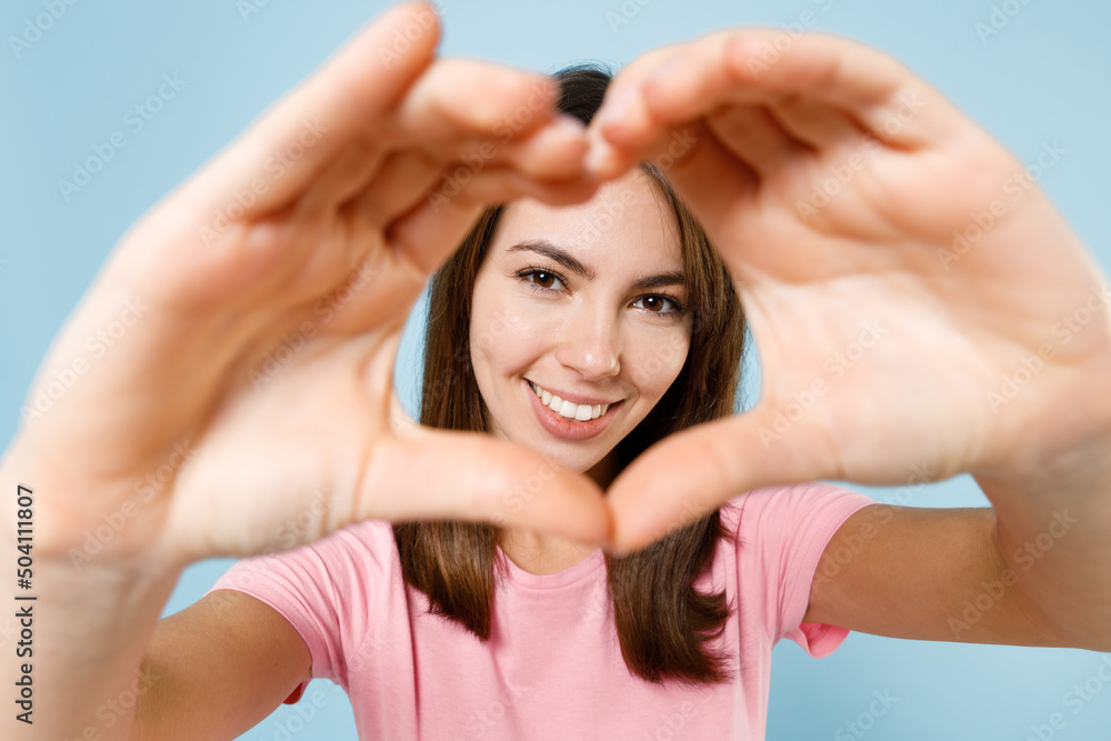 Close up young smiling happy woman 20s in pink t-shirt showing shape heart look through hands heart-shape sign isolated on pastel plain light blue background studio portrait. People lifestyle concept. Close up young smiling happy woman 20s in pink t-shirt showing shape heart look through hands heart-shape sign isolated on pastel plain light blue background studio portrait. People lifestyle concept.