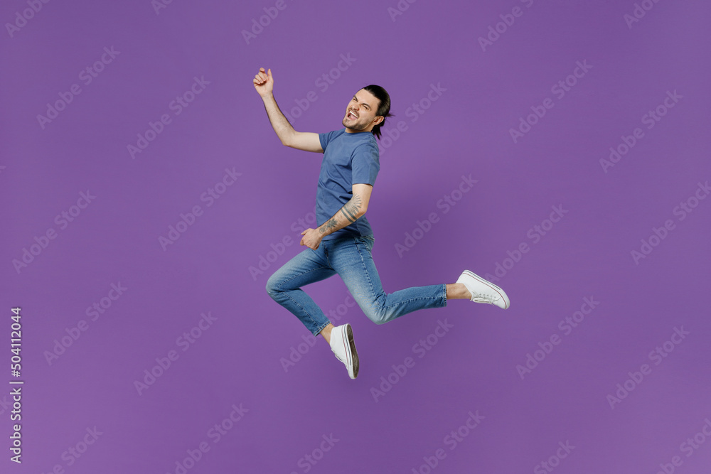 Full body young expressive singer rocker cool man 20s wearing basic blue t-shirt jump high do playing guitar gesture isolated on plain purple color background studio portrait People lifestyle concept