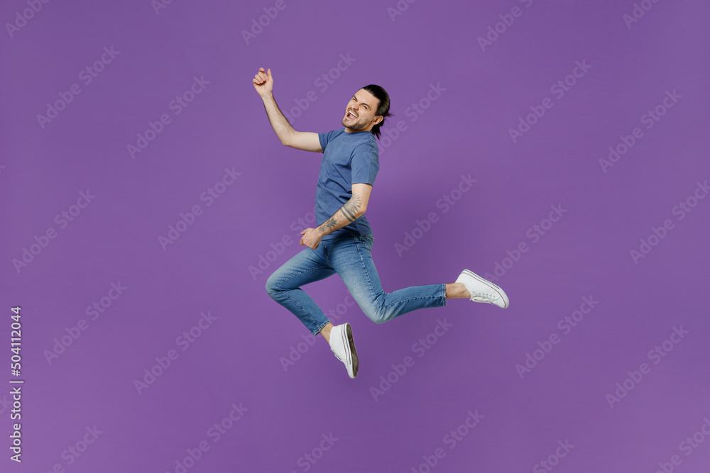 Full body young expressive singer rocker cool man 20s wearing basic blue t-shirt jump high do playing guitar gesture isolated on plain purple color background studio portrait People lifestyle concept Full body young expressive singer rocker cool man 20s wearing basic blue t-shirt jump high do playing guitar gesture isolated on plain purple color background studio portrait People lifestyle concept