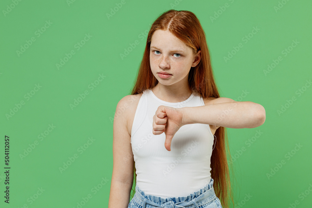 Little redhead kid disappointed sad girl 12-13 years old wearing white tank shirt showing thumb down dislike gesture isolated on plain green background studio portrait. Childhood lifestyle concept - obrazy, fototapety, plakaty 