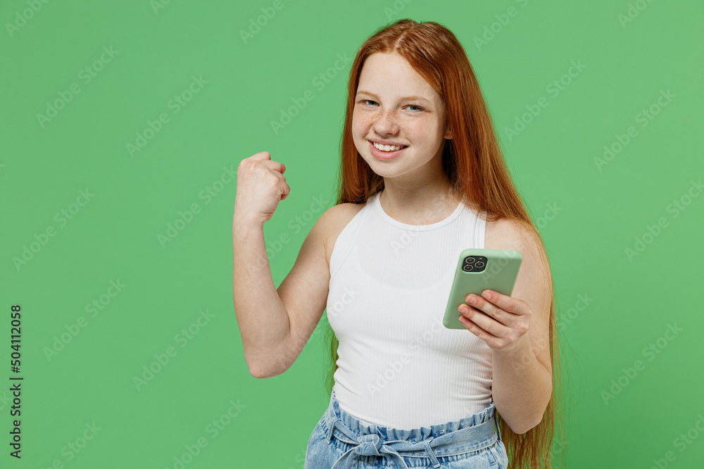 Little redhead kid ovejoyed girl 12-13 years old wearing white tank shirt hold in hand use mobile cell phone do winner gesture isolated on plain green background studio. Childhood lifestyle concept. Little redhead kid ovejoyed girl 12-13 years old wearing white tank shirt hold in hand use mobile cell phone do winner gesture isolated on plain green background studio. Childhood lifestyle concept.