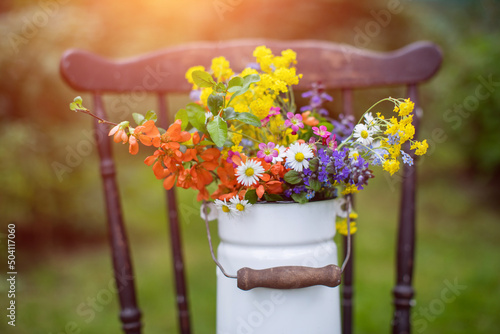 bukiet wiosennych, polnych kwiatów w rustykalnym klimaci w kance na mleko na starym krześle w ogrodzie w promieniach słońca, wiejskie klimaty