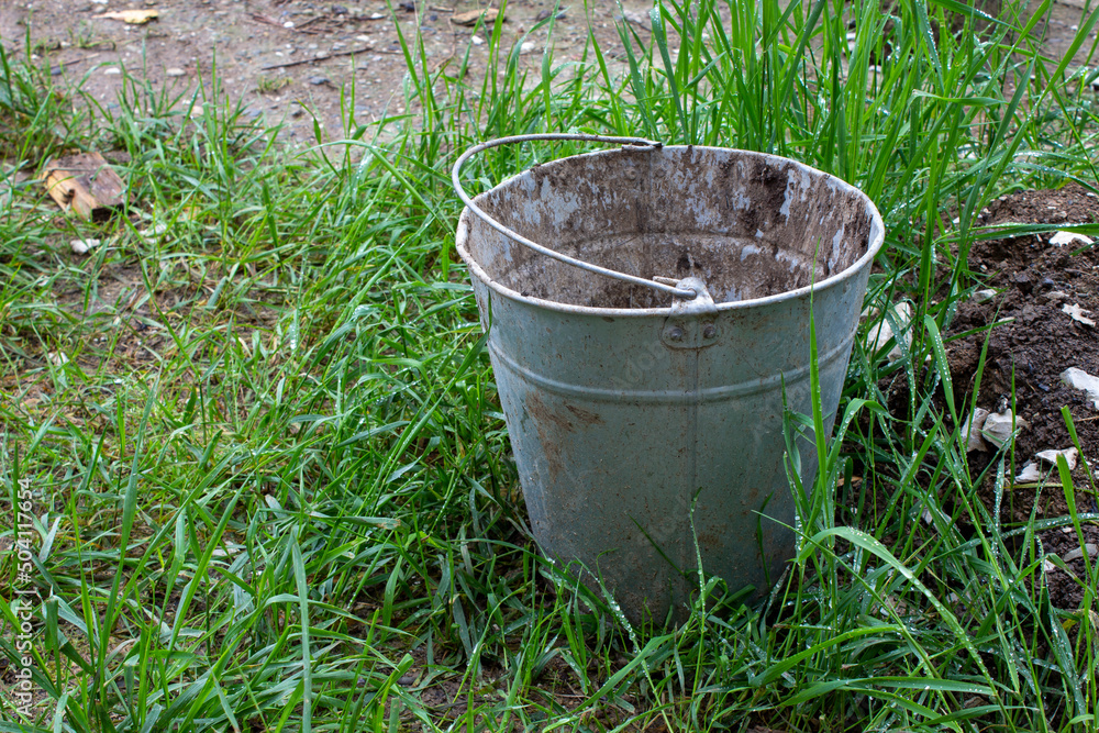 Old dirty empty bucket stained with paint on the street on the grass. Used metal bucket. Old farm. Old dirty empty bucket stained with paint on the street on the grass. Used metal bucket. Old farm.