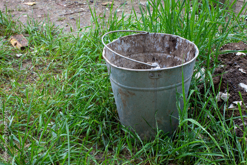 Old dirty empty bucket stained with paint on the street on the grass. Used metal bucket. Old farm.