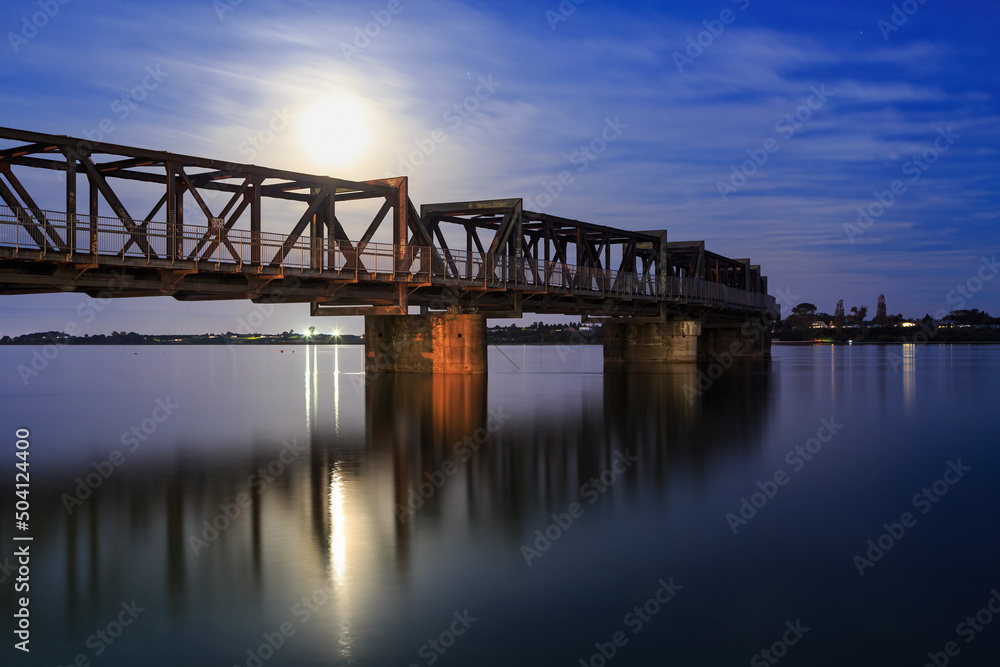 The full moon rising behind an old rail bridge. Both are reflected in the rippling water below. Photographed in Tauranga Harbour, New Zealand