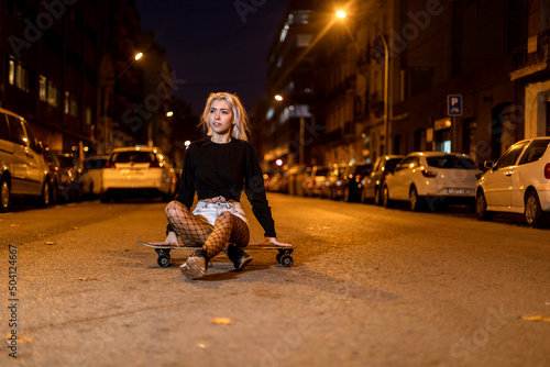 young woman in shorts on a long board while looking camera at night in the city