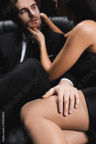 blurred man in suit touching leg of sexy woman on couch isolated on black.