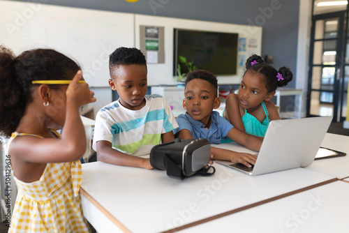 African american elementary school students using vr glasses and laptop at desk in classroom