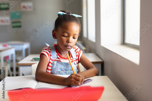 African american elementary schoolgirl writing on book while sitting at desk in classroom