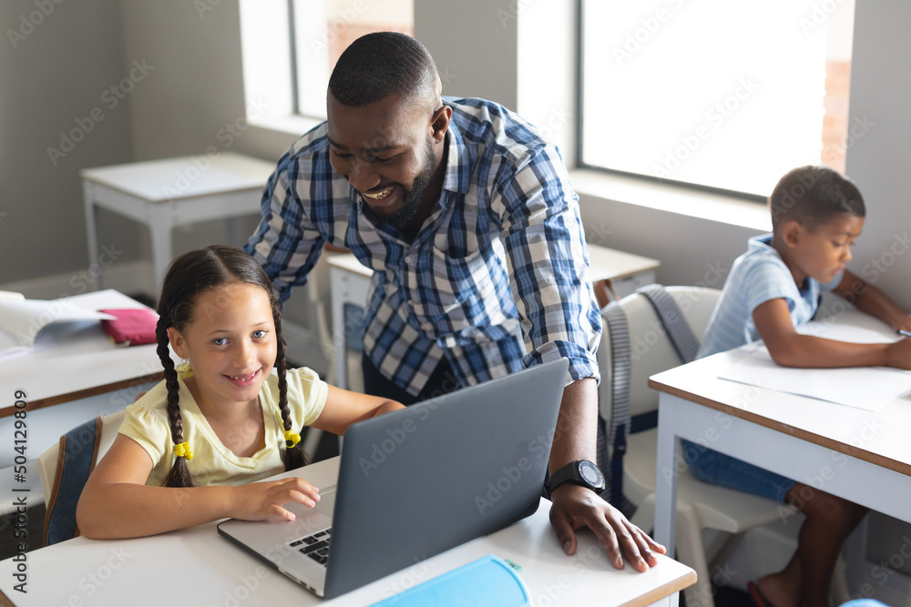 African american young male teacher looking at caucasian elementary schoolgirl with laptop African american young male teacher looking at caucasian elementary schoolgirl with laptop