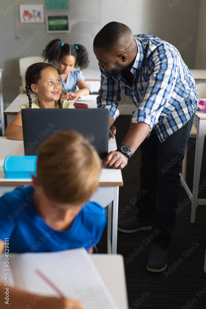 Smiling african american young male teacher looking at caucasian elementary schoolgirl with laptop Smiling african american young male teacher looking at caucasian elementary schoolgirl with laptop