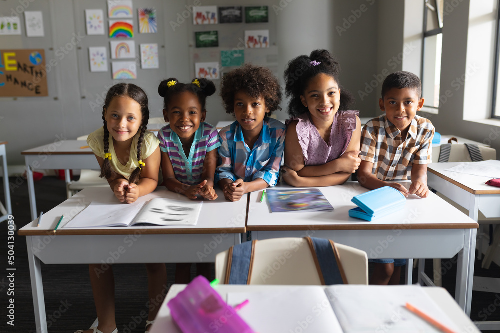 Smiling multiracial elementary school students sitting at desk in classroom - obrazy, fototapety, plakaty 