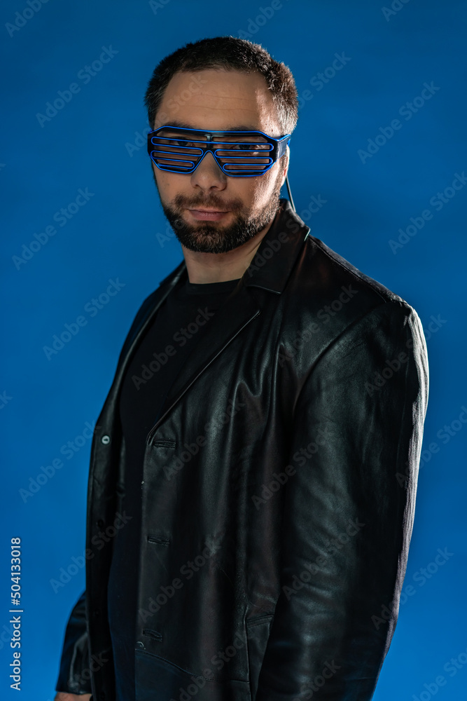 Obraz premium portrait of a man in interactive, cyber glasses. cyberpunk in the studio in black leather clothes. cool trendy handsome guy in flowy leather coat. bearded man with a gun, money on a blue background