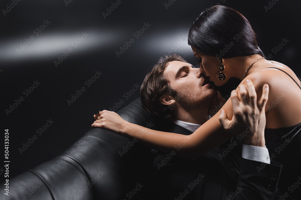 Young man in suit kissing brunette girlfriend on couch on black background.