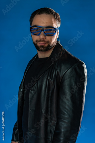 portrait of a man in interactive, cyber glasses. cyberpunk in the studio in black leather clothes. cool trendy handsome guy in flowy leather coat. bearded man with a gun, money on a blue background