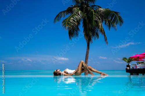 Elegant tanned woman in white swimsuit in pool on tropical Maldives island. Bikini girl in pool with view on horizon. Sexy model in the pool on beautiful Maldives landscape.Travel.