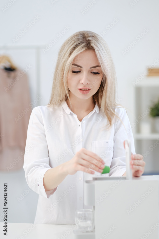 A young beautiful blonde woman in a white shirt brews coffee in capsule coffee machine. Bright home interior. A young beautiful blonde woman in a white shirt brews coffee in capsule coffee machine. Bright home interior.
