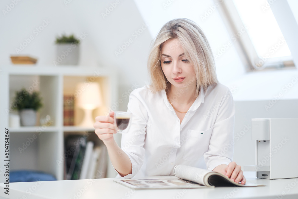 A young beautiful Caucasian blonde woman rests at home. A woman sits at a table, drinking coffee from a glass cup and reading a magazine. A young beautiful Caucasian blonde woman rests at home. A woman sits at a table, drinking coffee from a glass cup and reading a magazine.