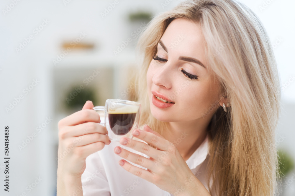 A young beautiful Caucasian blonde woman rests at home. A woman sits at a table, drinking coffee from a glass cup and reading a magazine. A young beautiful Caucasian blonde woman rests at home. A woman sits at a table, drinking coffee from a glass cup and reading a magazine.