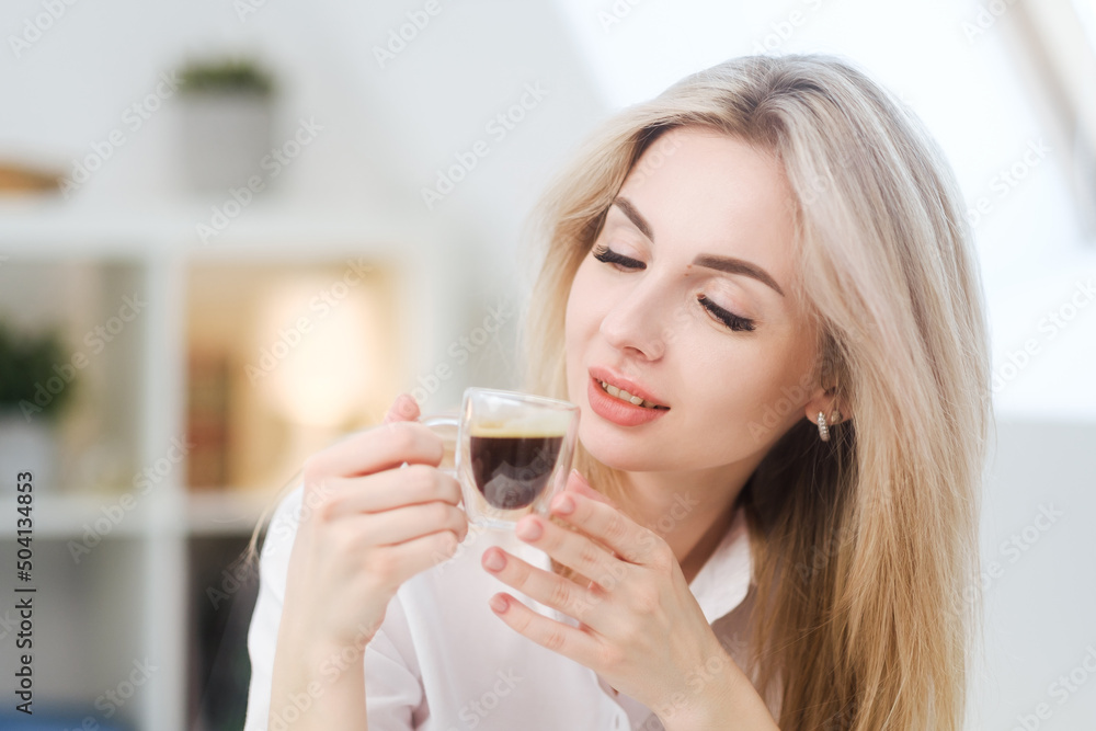 A young beautiful Caucasian blonde woman rests at home. A woman sits at a table, drinking coffee from a glass cup and reading a magazine. A young beautiful Caucasian blonde woman rests at home. A woman sits at a table, drinking coffee from a glass cup and reading a magazine.