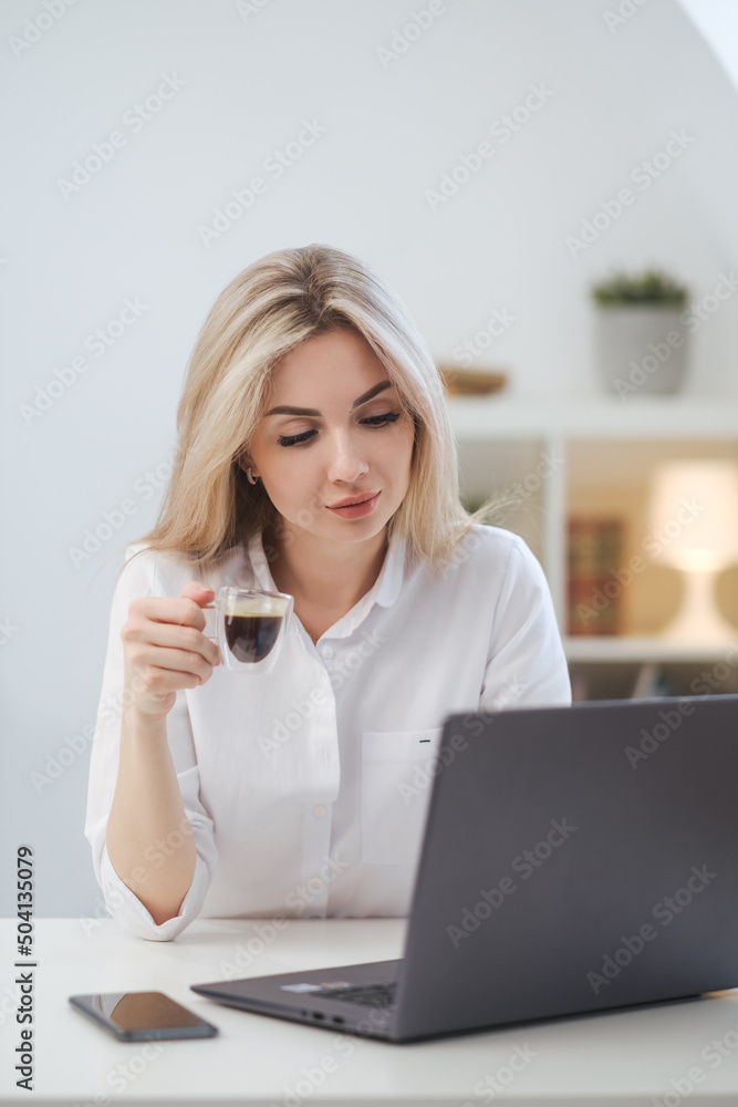 Young beautiful caucasian blonde woman working on laptop at home and drinking coffee. Remote work or freelance. Young beautiful caucasian blonde woman working on laptop at home and drinking coffee. Remote work or freelance.