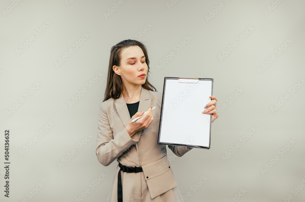 Business woman with a tablet in her hands stands on a beige background looks at a blank white sheet of paper and shows a pen on copy space Business woman with a tablet in her hands stands on a beige background looks at a blank white sheet of paper and shows a pen on copy space