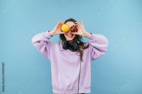 Happy girl in purple sweatshirt raised apples to eye level on blue background and smiles. © bodnarphoto