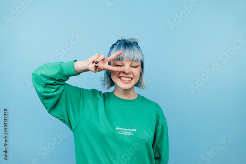 Obraz na plátne Attractive hipster girl in casual clothes stands on a blue background and shows a gesture of peace winking