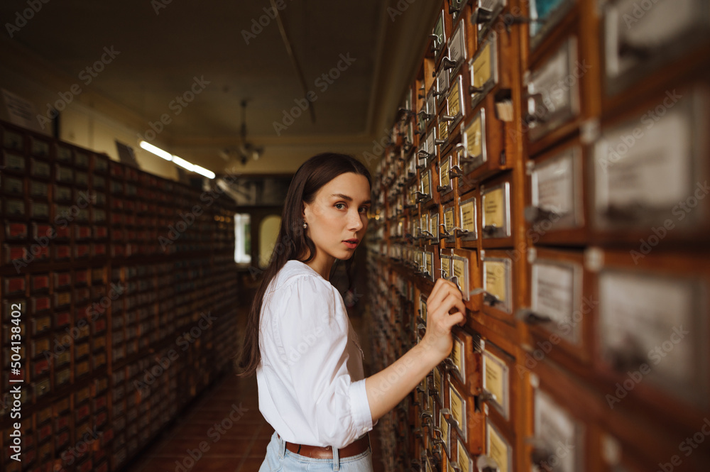 Beautiful woman in a white blouse stands in the archives of the public library, opens a bedside table in search of information and looks at the camera. Beautiful woman in a white blouse stands in the archives of the public library, opens a bedside table in search of information and looks at the camera.