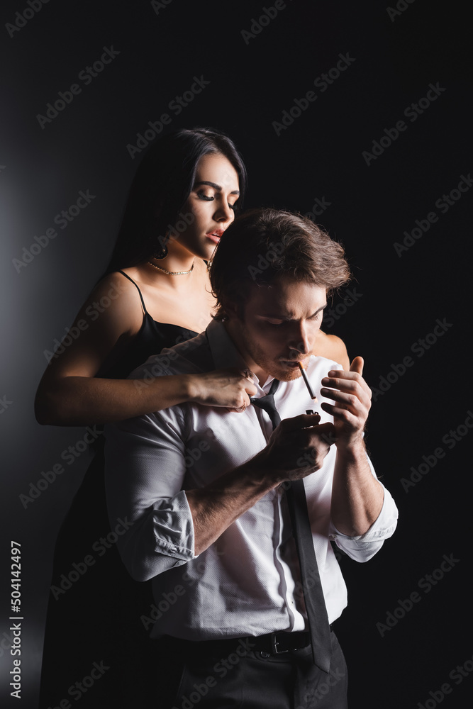 Sexy woman undressing boyfriend in formal wear smoking on black background.