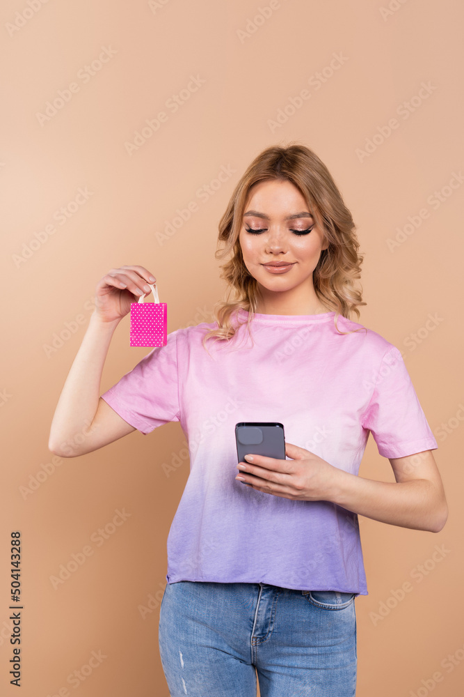 pleased woman holding smartphone and tiny shopping bag isolated on beige. pleased woman holding smartphone and tiny shopping bag isolated on beige.
