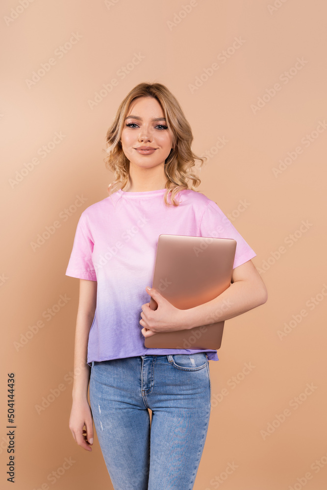 happy woman with wavy hair looking at camera while holding laptop isolated on beige. happy woman with wavy hair looking at camera while holding laptop isolated on beige.