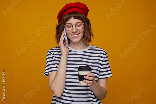 indoor portrait of young ginger female, wears stripped t shirt and glasses posing over orange background holding cup of coffee and talking on phone