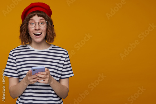 indoor portrait of young ginger female, wears stripped t shirt and glasses posing over orange background excited, surprised facial expression