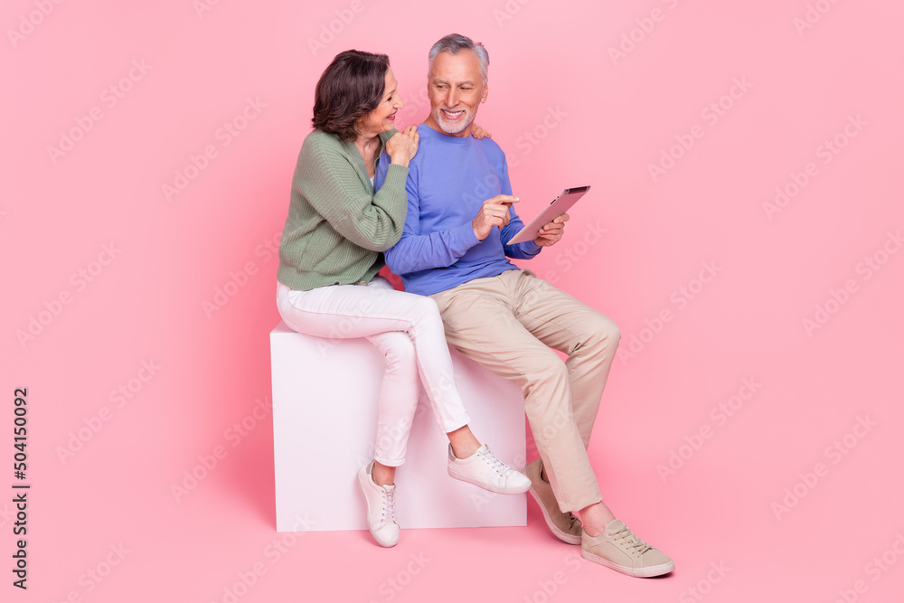 Full length body size view of two attractive cheery people sitting on box using device isolated over pink pastel color background Full length body size view of two attractive cheery people sitting on box using device isolated over pink pastel color background