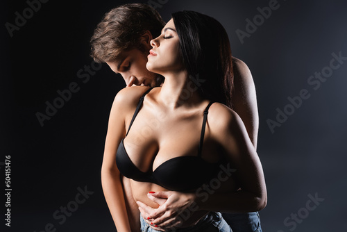 Side view of shirtless man kissing shoulder of sexy woman on black background.
